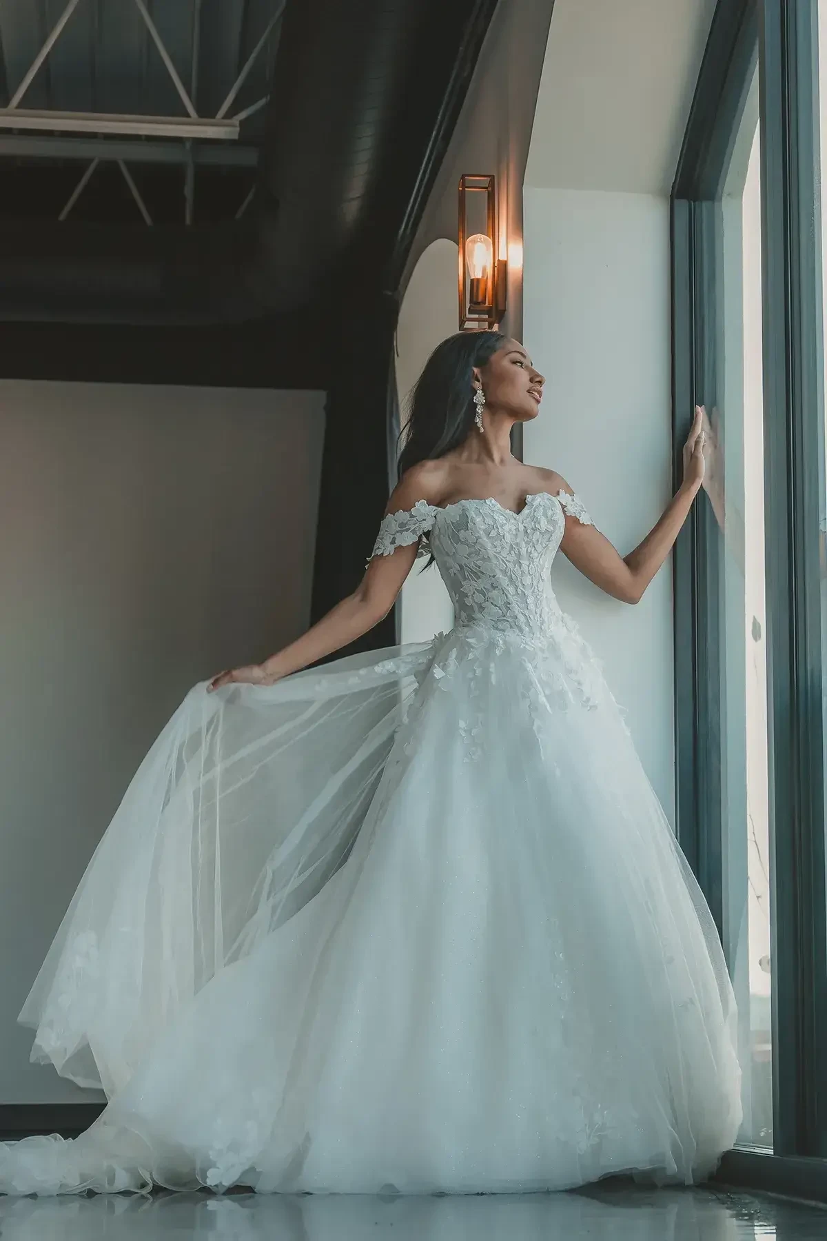 A woman in a beautiful white wedding dress stands by a large window, gazing thoughtfully outside. The dress features off-the-shoulder sleeves and a flowing train.