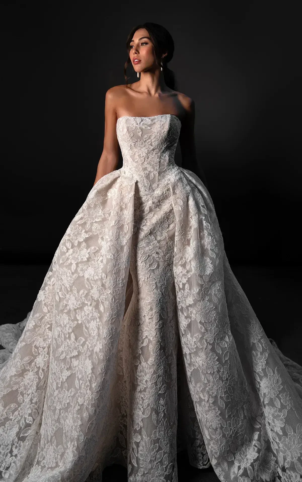 A woman wearing a strapless, intricately designed lace gown with a voluminous skirt, standing against a dark background.