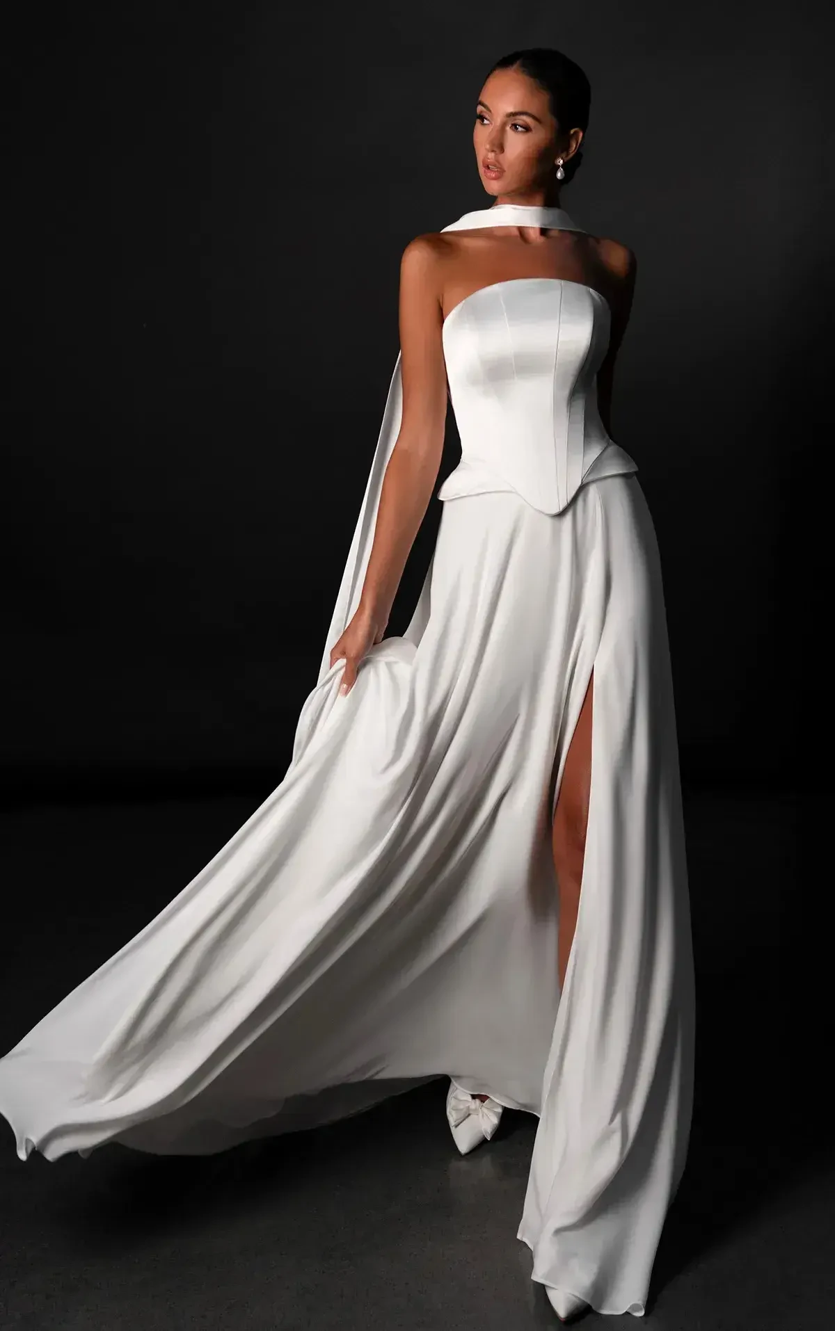 A model wearing an elegant white gown with a high slit and a stylized neckline, posed against a dark background.